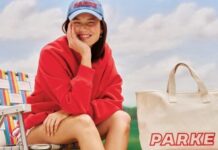 Target Launches Parke Collab With Apparel, Denim and Swim Smiling woman in a red sweatshirt and blue cap sits on a striped beach chair outdoors beside a beige PARKE tote bag.