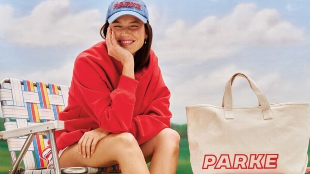 Smiling woman in a red sweatshirt and blue cap sits on a striped beach chair outdoors beside a beige PARKE tote bag.