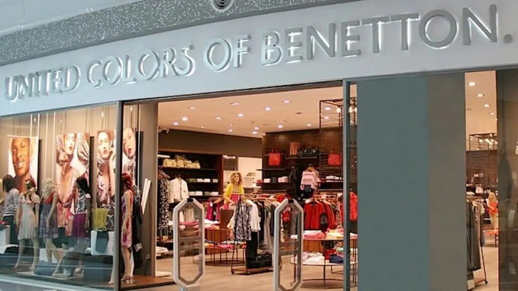 Storefront of a United Colors of Benetton shop in a mall, with mannequins and clothing displays inside the brightly lit store sign visible above.
