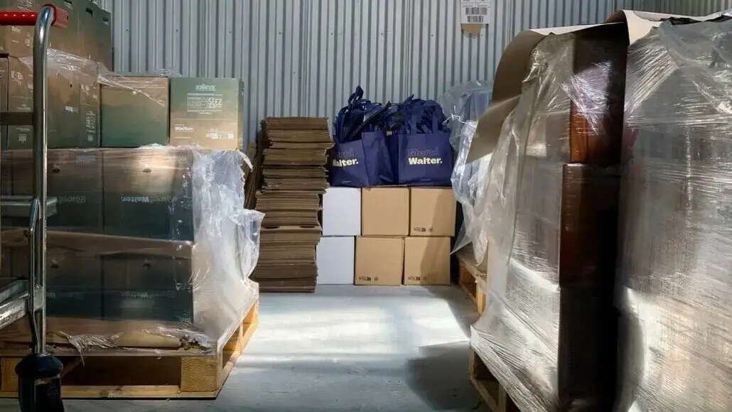 Warehouse storage area with stacked boxes on pallets, plastic wrap, and blue Walter bags in the center.