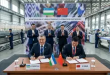 Uzbekistan Textile Automation Memorandum Signed with Chinese Partner Two men sign documents at a table in a textile factory, Uzbekistan and China flags between them as officials applaud behind them in a formal ceremony on a banner backdrop.