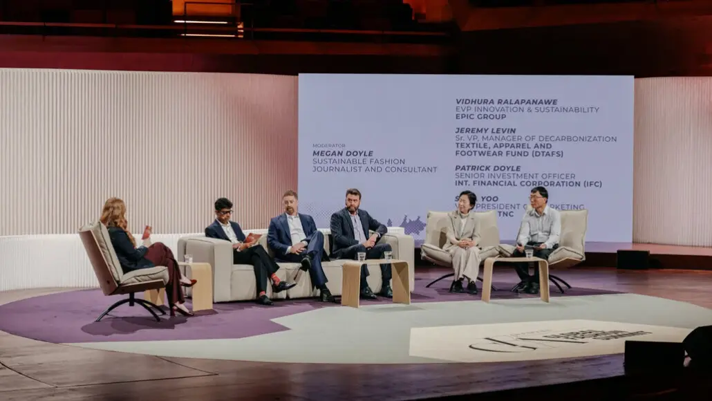 Panel discussion on a stage with six people seated in white chairs, moderator on the left, and a large screen displaying speakers' names in the background.