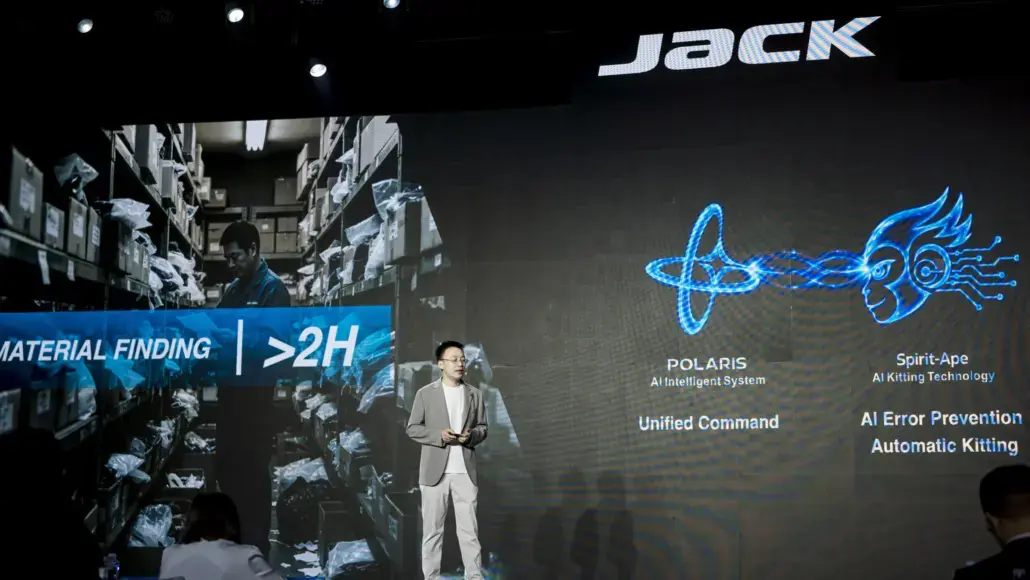 Presenter on a stage giving a talk, with a large display showing a warehouse scene and AI/kitting technology labels such as 'MATERIAL FINDING >2H' and logos 'POLARIS' and 'Spirit-Ape' on the screen.