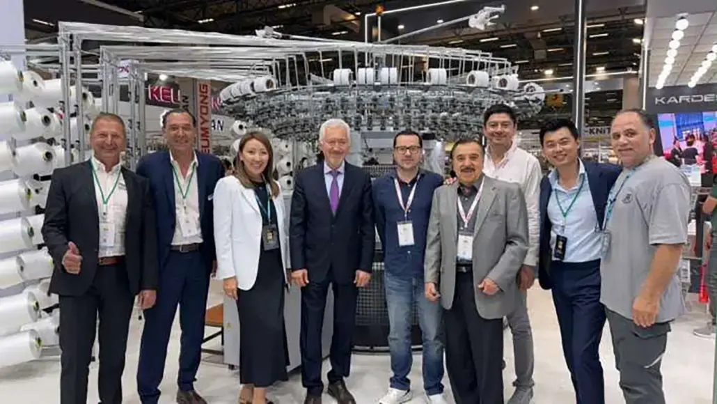 Ten professionals in business attire pose in front of a large industrial textile machine at an exhibition booth, badges visible around their necks.