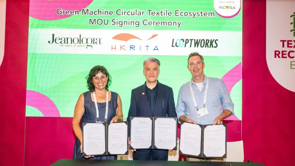 Three adults stand behind a table, each holding an open certificate during a signing ceremony with a green and pink backdrop.