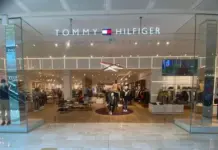 PVH Cuts 2026 Revenue Outlook as EMEA Demand Softens Tommy Hilfiger storefront with glass doors, bright lighting, and clothing displays inside.