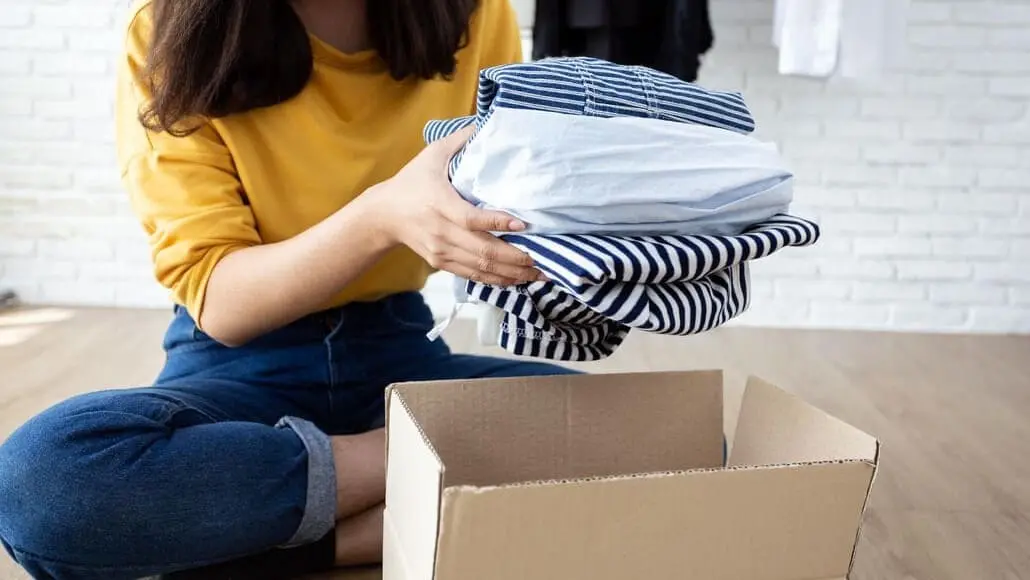 Person folding striped shirts and placing them into an open cardboard box on the floor there is a light brick wall in the background and the person wears a yellow top and blue jeans