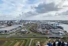 Dutch Industrial Production Rises in April Amidst Shifting Producer Sentiment View of a busy industrial port with container yard, cranes, warehouses, and railroad tracks; people watching from a high vantage point in the foreground.