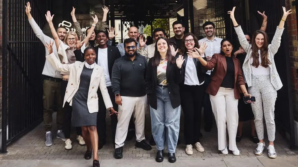 Group of diverse colleagues posing outside a building, smiling and raising their hands in celebration. A cheerful, mixed-gender team photo taken at an office entrance with hands raised. A large team of coworkers posing for a group photo, showing unity and enthusiasm. A diverse group of professionals standing on a sidewalk, arms in the air, celebrating together.