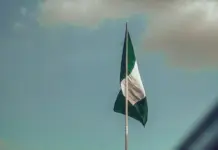 Manufacturers Urge Dialogue on Proposed Textile Import Ban in Nigeria Nigerian flag waving on a tall flagpole against a blue sky with clouds above.