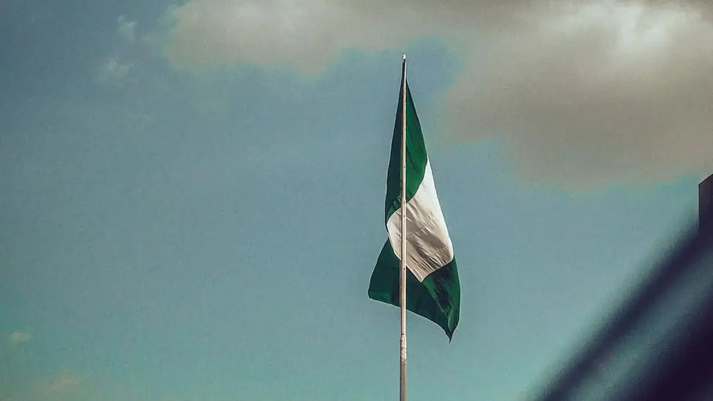 Nigerian flag waving on a tall flagpole against a blue sky with clouds above.