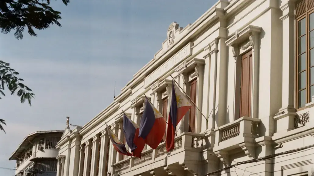 White neoclassical building with ornate balconies and several hanging flags against a clear sky
