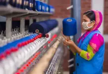 Report: Downward T-Shirt Pricing Hurts Garment Wages and Safety A woman wearing a colorful outfit and mask inspects a textile loom with blue yarn spools nearby