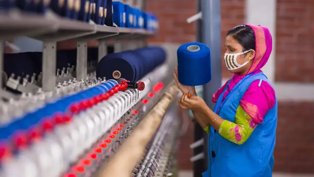 A woman wearing a colorful outfit and mask inspects a textile loom with blue yarn spools nearby