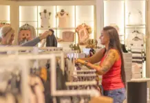 UK Retail Sales Rise in May as Heatwave Lifts Clothing Two women in a clothing store browsing racks; one in a red tank top shows a garment to the other, who smiles and holds up a piece of clothing.