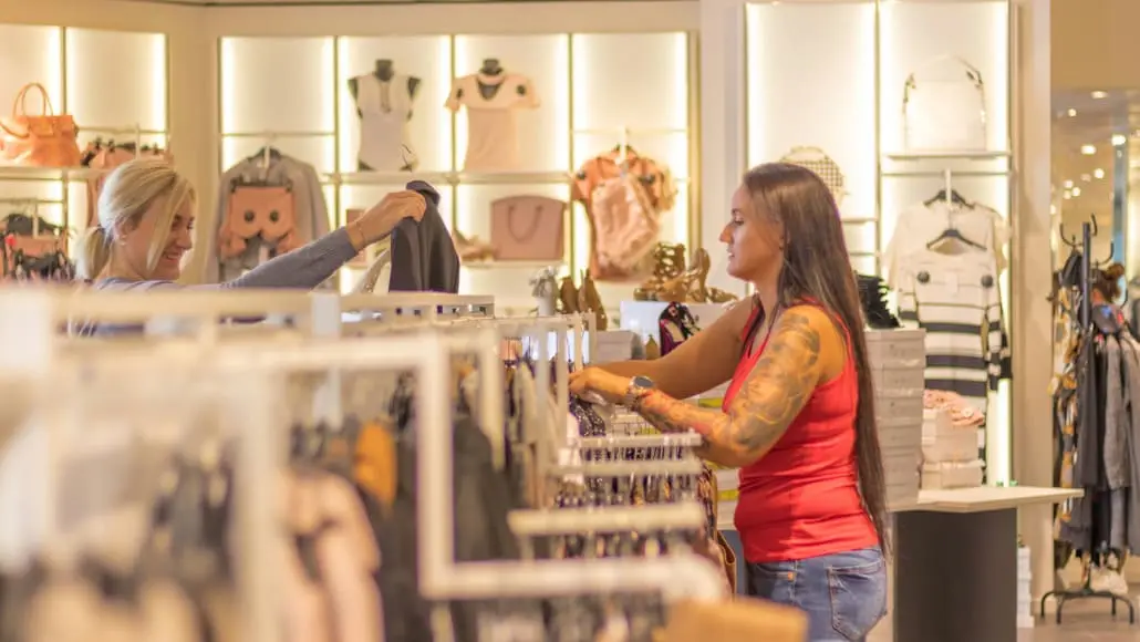 Two women in a clothing store browsing racks; one in a red tank top shows a garment to the other, who smiles and holds up a piece of clothing.