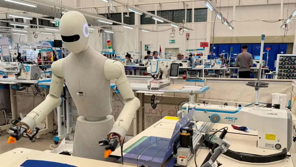 Life-size humanoid robot standing at a sewing workstation in a bright workshop, with sewing machines and workers in the background.