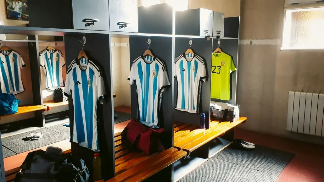 Locker room with light-blue and white striped soccer jerseys hanging in individual stalls, benches with gear nearby.