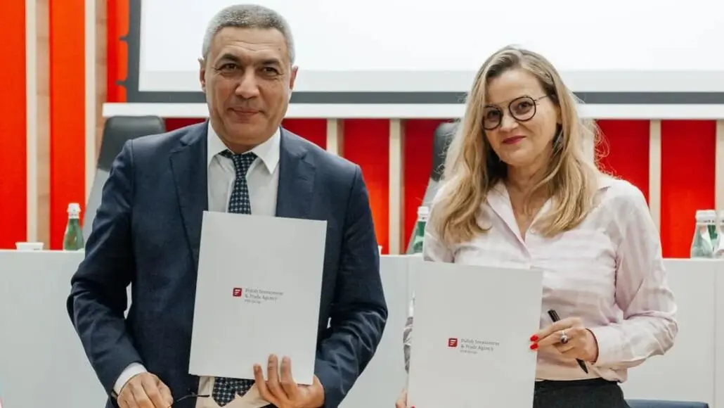 Two professionals in a conference room pose with signed documents in front of a red and white backdrop, facing the camera.