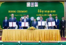 Germany and GIZ Support Sustainable Textile Industry in Cambodia Officials sign documents at a ceremony, standing behind a long yellow-clothed table with a green backdrop that reads 'SIGNING CEREMONY' and flags on both sides.