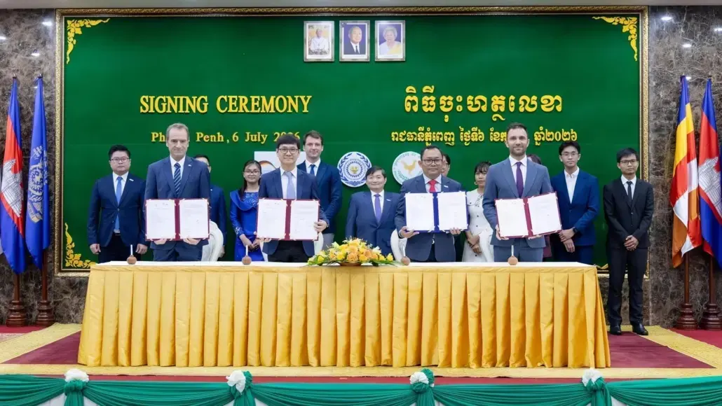 Officials sign documents at a ceremony, standing behind a long yellow-clothed table with a green backdrop that reads 'SIGNING CEREMONY' and flags on both sides.