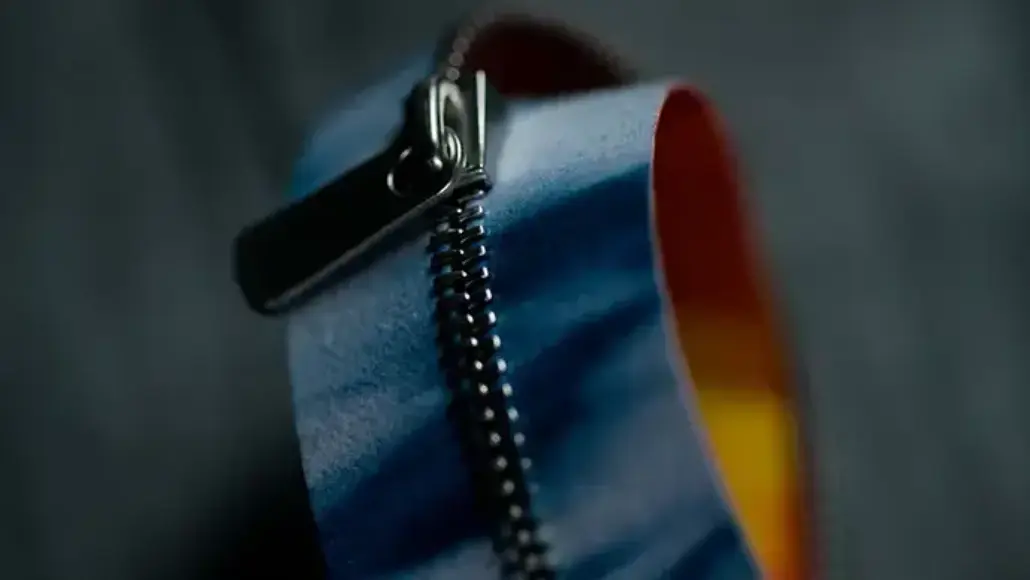 Close-up of a metal zipper slider on blue fabric with a rainbow-colored edge.