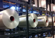 Cygnet Texkimp Develops 500kg VHD PAN Precursor Unwinding Creel to Increase Efficiency in Carbon Fibre Production Row of large silver rolls with red cores on a metal conveyor rack in a factory setting.