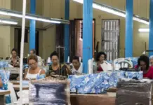 Lesotho Regains Duty-Free Export Status Following Expiration of United States Tariffs People seated at sewing machines in a textile workshop, working on blue-patterned fabric under bright fluorescent lights with blue columns nearby.