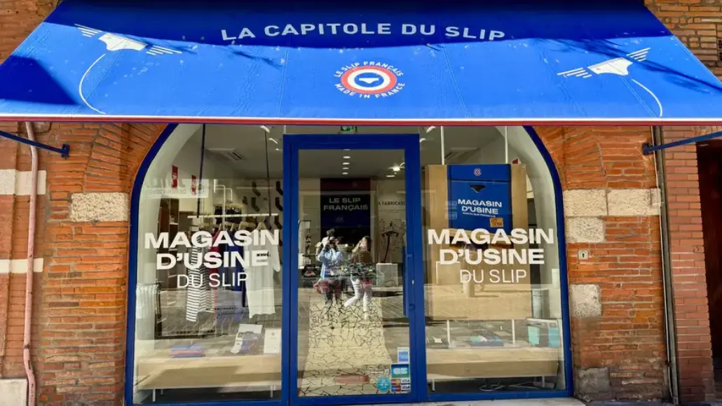 Blue awning storefront for La Capitole du Slip, brick facade and glass doors with 'MAGASIN D'USINE DU SLIP' text on the windows/doors as a factory outlet shop sign.