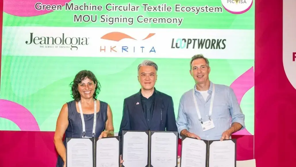 Three people holding signed MOUs at the Green Machine Circular Textile Ecosystem signing ceremony with a bright green backdrop beside sponsor logos