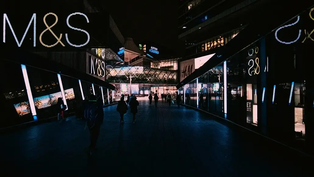 Nighttime pedestrian street lined with illuminated M&S storefronts and shoppers walking by.
