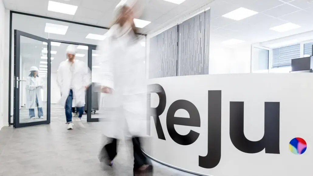 Researchers in white lab coats walk through a bright, modern reception area with a curved desk displaying the word 'Relj' and a small colorful logo.