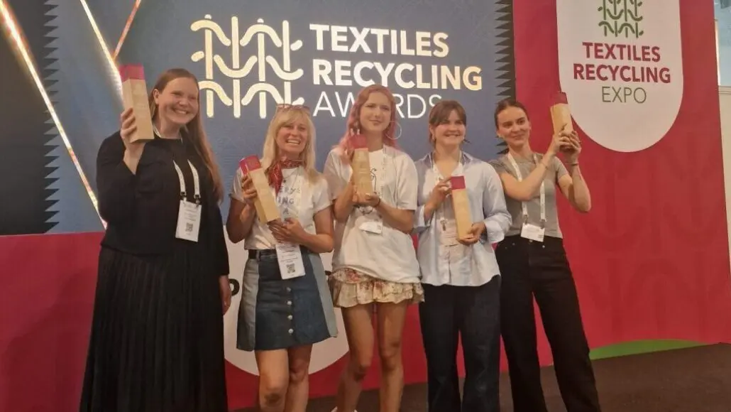 Six women posing on a stage at a Textiles Recycling Awards event, each holding a tall wooden trophy in front of a blue and red backdrop reading 'Textiles Recycling Awards'.