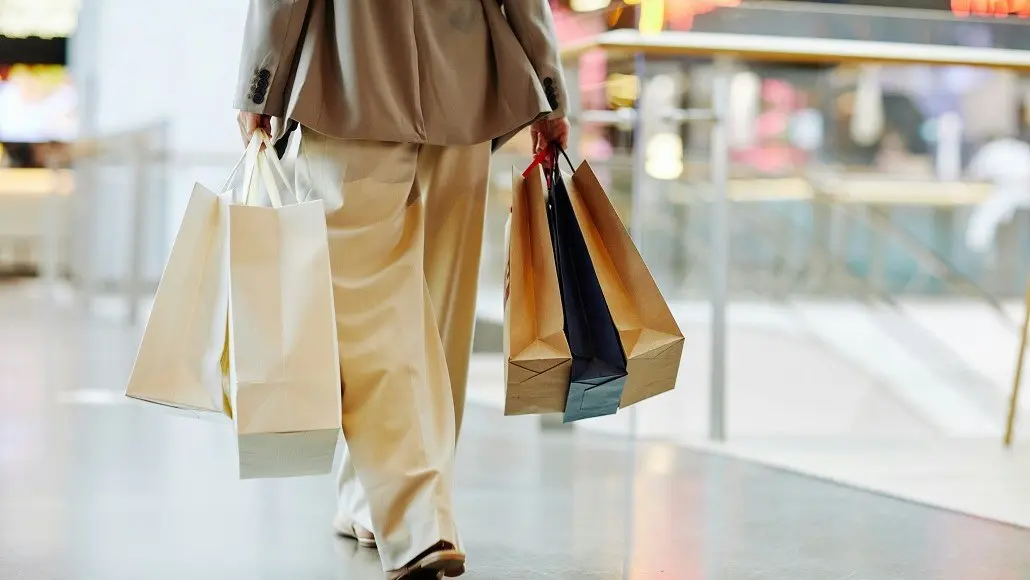Person in a tan suit walks through a shopping mall carrying multiple shopping bags in each hand.