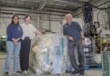 Goodwill, Ontario Universities Advance Textile Recycling with AI Three people in a warehouse pose beside a large wrapped bale of recycled paper near industrial equipment.