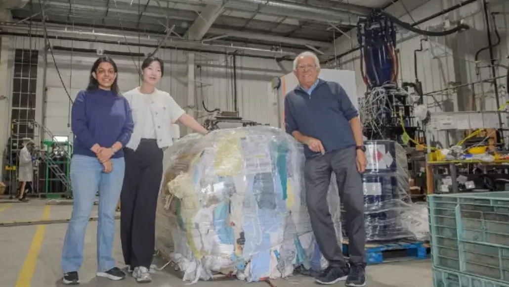 Three people in a warehouse pose beside a large wrapped bale of recycled paper near industrial equipment.
