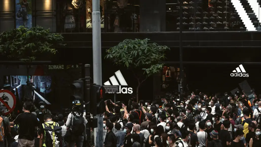 Crowd of people wearing masks gathered outside an Adidas storefront with large logo signs.