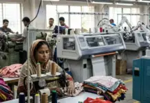 Automation Threatens Over One Million Garment Jobs in Bangladesh Woman sewing fabric on a vintage machine in a busy textile factory, with colorful cloth piles and other workers in the background.