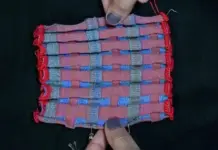 Harvard Researchers Develop Innovative Programmable Knitted Textiles Close-up of a pink and blue woven fabric swatch with red edging, held by fingers against a dark background.