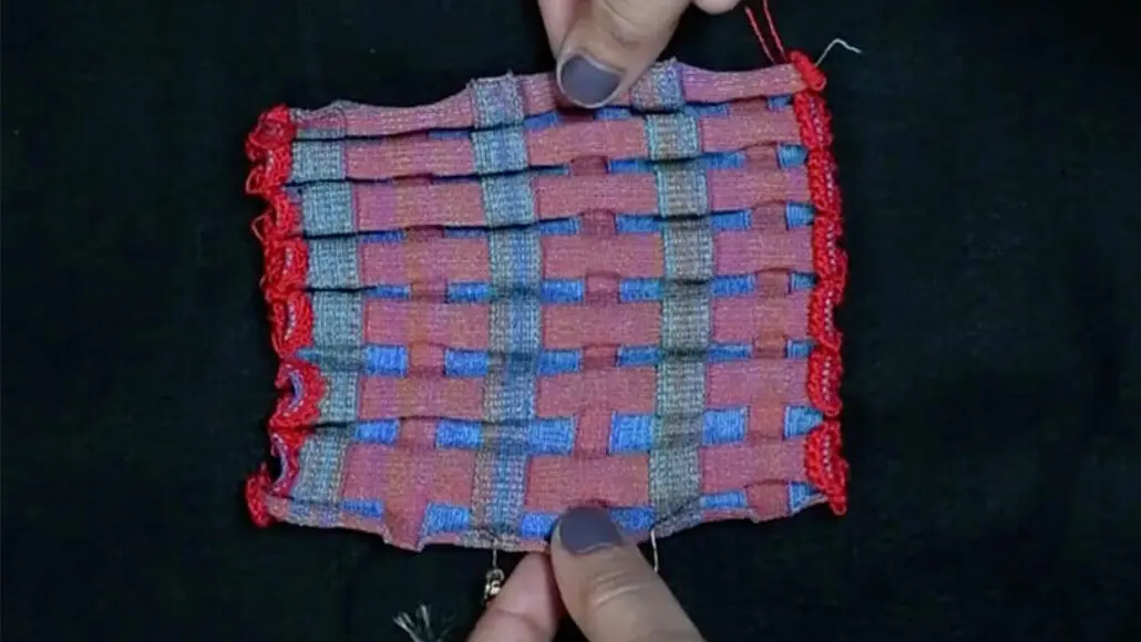 Close-up of a pink and blue woven fabric swatch with red edging, held by fingers against a dark background.