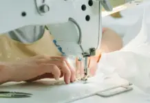 EU Launches Digital Product Passport Registry as Textile Ecodesign Work Advances Close-up of hands guiding white fabric under a sewing machine needle during stitching in an industrial setup.