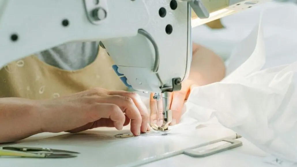 Close-up of hands guiding white fabric under a sewing machine needle during stitching in an industrial setup.