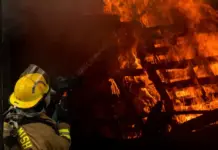 Fatal Fire at Jinjiang Shoe Factory Claims 28 Lives Firefighter in yellow helmet and turnout gear directs a hose at a large blaze consuming stacked wooden pallets temporarily obscured by smoke and darkness.