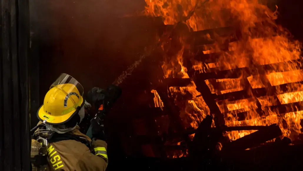 Firefighter in yellow helmet and turnout gear directs a hose at a large blaze consuming stacked wooden pallets temporarily obscured by smoke and darkness.