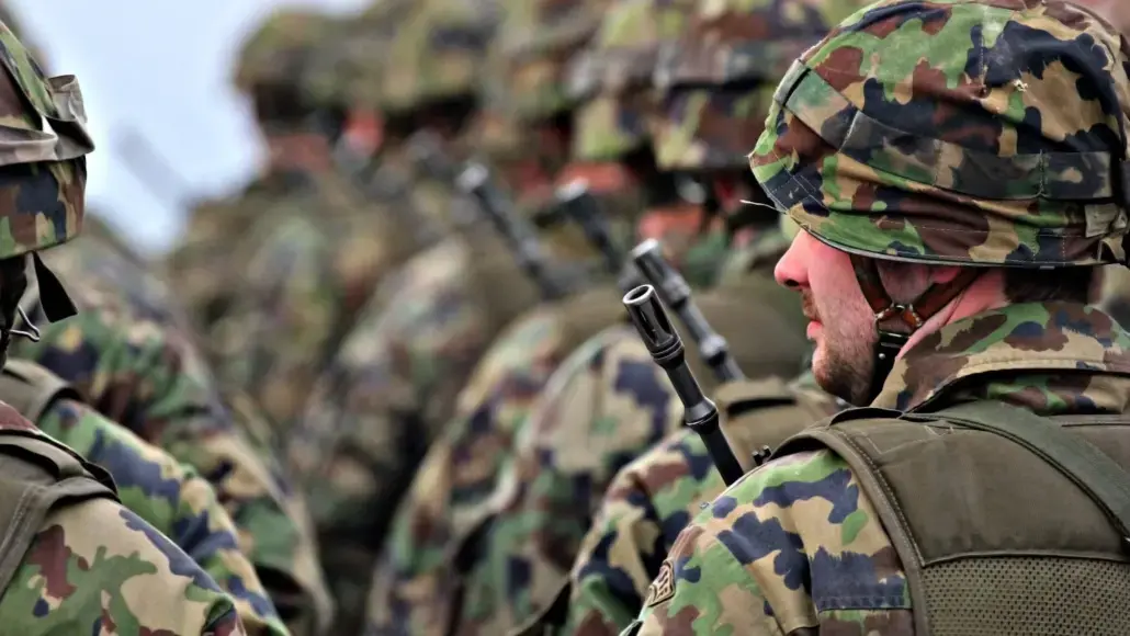 Soldier in camouflage uniform and helmet with a rifle, standing among other soldiers in a field formation.