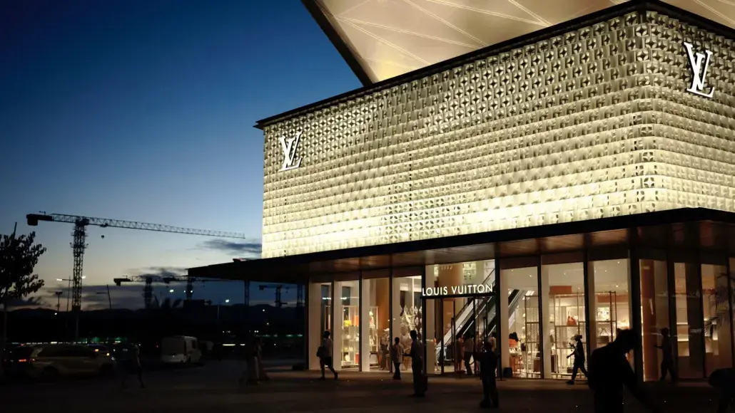 Louis Vuitton storefront at dusk with a lit patterned façade and shoppers entering the glass doors.