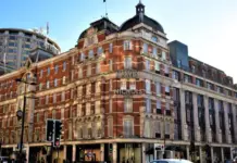 Next Reportedly Weighs Harvey Nichols Takeover to Expand Luxury Retail Presence Red brick Harvey Nichols department store on a busy city street with classic arched windows and a curved corner facade in central London/near Piccadilly Circus area