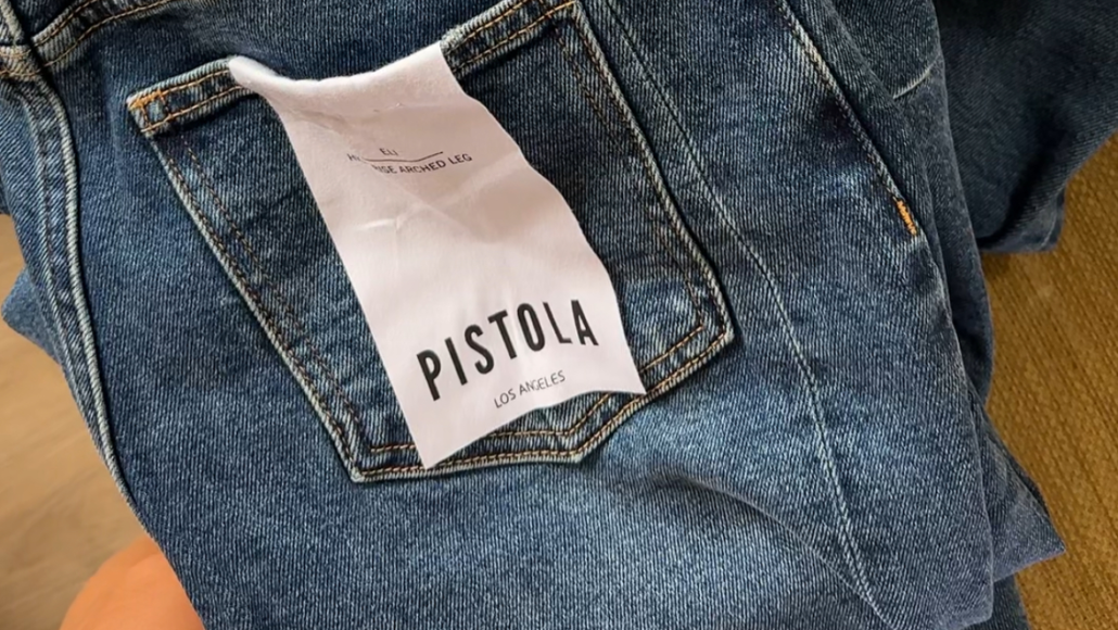Back pocket of blue jeans with a white tag reading 'PISTOLA' and 'LOS ANGELES'