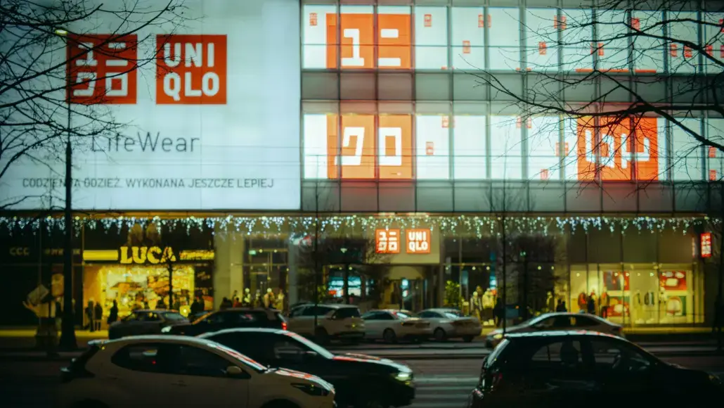 UNIQLO storefront at night with large orange logo, LifeWear banner, and string lights; busy street with cars in foreground.