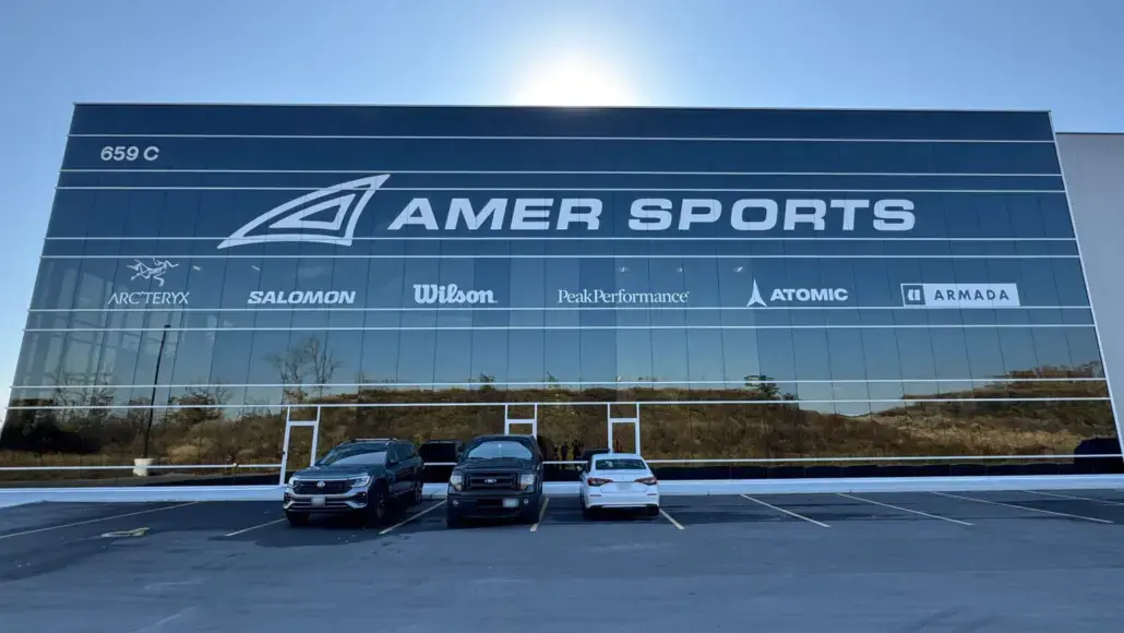 Facade of the Amer Sports building with a large logo and brand logos (Arc'teryx, Salomon, Wilson, PeakPerformance, Atomic, Armada) and three cars parked in front.